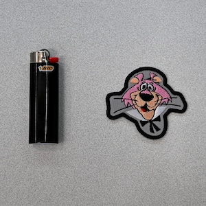 May include: A black lighter with a yellow "BIC" logo next to a gray and pink embroidered patch of a cartoon cat wearing a bow tie.