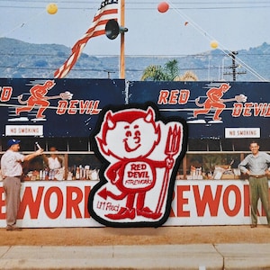 May include: Vintage photograph of a fireworks stand with the text "FIREWORKS" in large red letters. A patch of the Red Devil Fireworks logo is in the foreground. The American flag waves in the background.