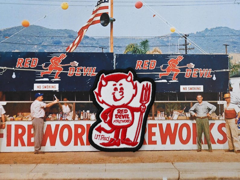 May include: Vintage photograph of a fireworks stand with the text "FIREWORKS" in large red letters. A patch of the Red Devil Fireworks logo is in the foreground. The American flag waves in the background.