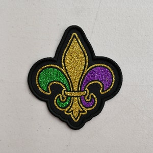May include: A black felt patch with a gold, green, and purple glitter fleur-de-lis design.