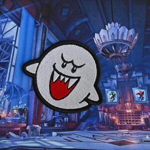 Puede incluir: Un fantasma blanco con una sonrisa roja y dientes, bordado en un parche de tela. El fantasma est&aacute; en una habitaci&oacute;n oscura de estilo g&oacute;tico con una l&aacute;mpara de ara&ntilde;a y otros elementos decorativos.