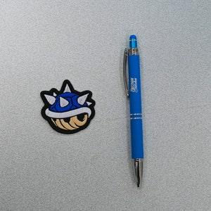 May include: A blue and white embroidered patch of a spiky, shelled character from the Super Mario Bros. video game series. The character is known as a Spiny Shell.