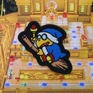 May include: Embroidered patch featuring a cartoon character in a blue robe and hat, riding a broom. The character has yellow skin, glasses, and a red gem on a staff. The patch is on a game board background.