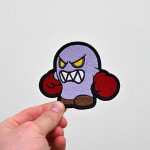 May include: Embroidered patch of a cartoon ghost character. The ghost is purple with red boxing gloves and brown feet. It has an angry expression with yellow eyes and a white mouth. The patch is outlined in black.