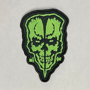 May include: Green embroidered skull patch with a black outline. The skull has a detailed design with a pointed chin and two small horns.
