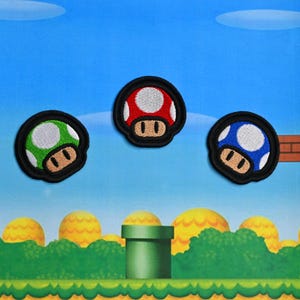 May include: Three embroidered mushroom patches in red, green, and blue with white polka dots and brown details. Each patch has a black border. The patches are set against a background reminiscent of a video game level.