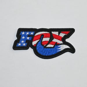 May include: Embroidered patch featuring the word "FOX" in a stylised font. The "O" is shaped like a fox tail and filled with an American flag design. The patch is black, red, white and blue.