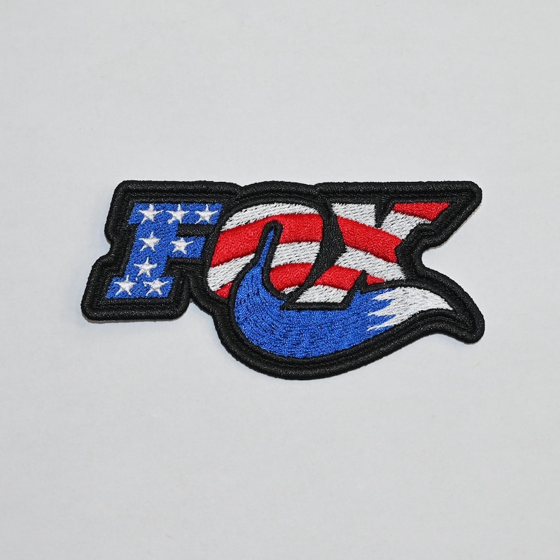 May include: Embroidered patch featuring the word "FOX" in a stylised font. The "O" is shaped like a fox tail and filled with an American flag design. The patch is black, red, white and blue.