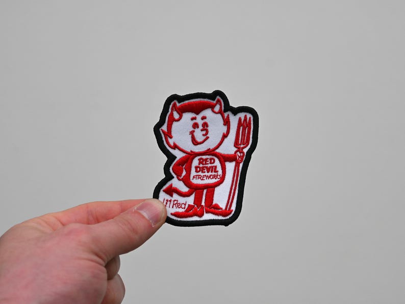 May include: Embroidered patch with a cartoon devil holding a pitchfork. The devil is red and white with a smiling face. The patch has the text "RED DEVIL FIREWORKS" and a black border.