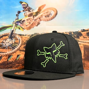 May include: Black baseball cap featuring a neon green Fox Racing logo with a skull and crossbones design. The cap has a flat brim and a structured fit. The background shows a motocross rider in action, mid-air on a dirt bike.