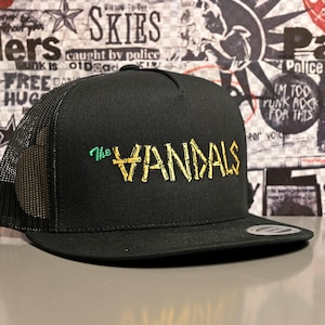 May include: Black trucker hat with a mesh back and a flat brim. The front of the hat features the text "The VANDALS" in gold and green embroidery. The hat is a classic style.