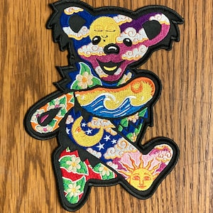 May include: A colorful embroidered patch featuring a cartoon bear with a psychedelic design. The bear is surrounded by clouds, stars, suns, and moons. The patch is perfect for adding a unique touch to clothing, bags, or other accessories.