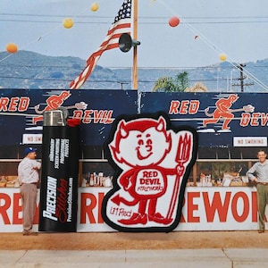 May include: A black lighter with "Precision Stitches" printed on it stands next to a red and white patch featuring a cartoon devil holding a pitchfork. The background includes a sign that says "Red Devil Fireworks".