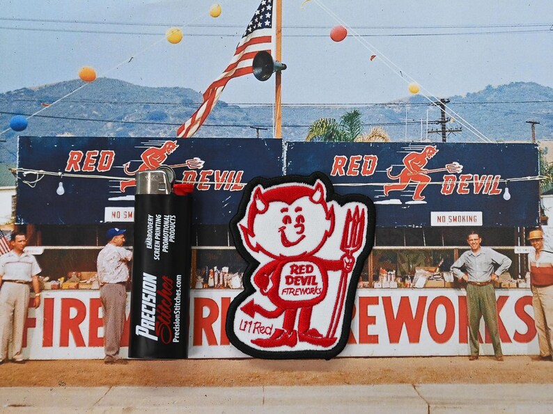 May include: A black lighter with "Precision Stitches" printed on it stands next to a red and white patch featuring a cartoon devil holding a pitchfork. The background includes a sign that says "Red Devil Fireworks".