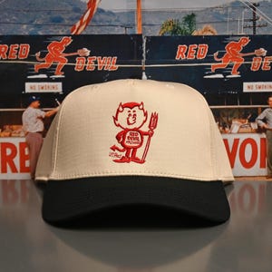 May include: A cream-colored baseball cap with a black brim. The cap features a red embroidered cartoon devil holding a pitchfork, with the text "Red Devil Archives" below. The background includes a vintage sign.