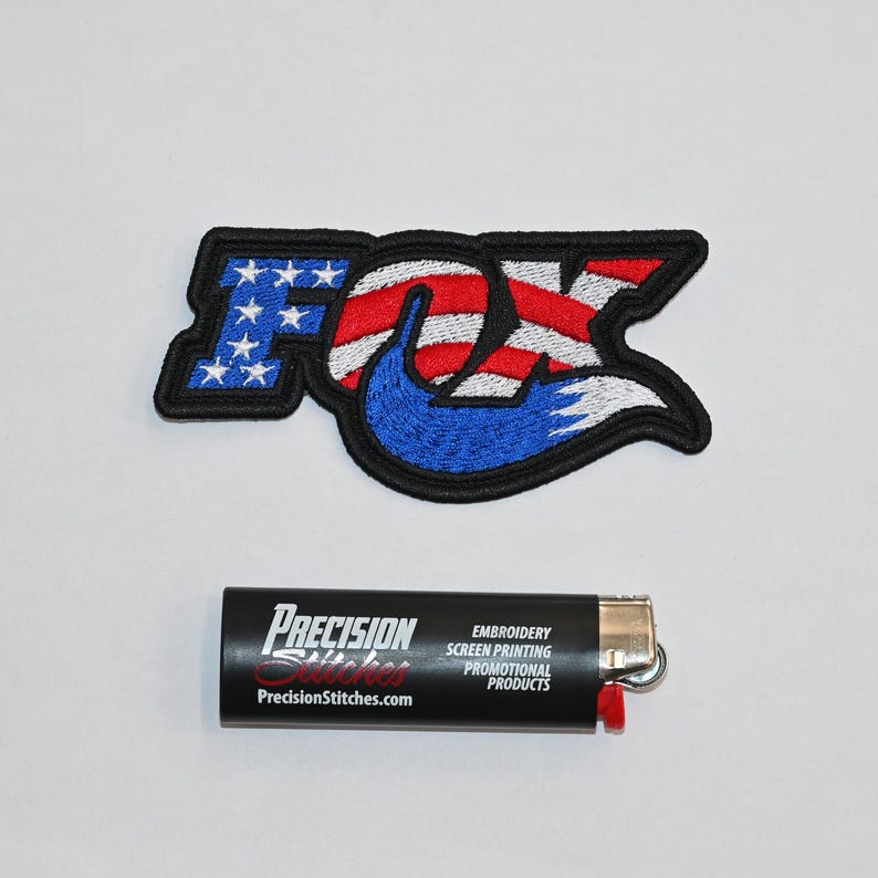 May include: An embroidered patch featuring the Fox logo with an American flag design. The patch is black with red, white, and blue stitching. The text "Precision Stitches.com" is visible on a black lighter.