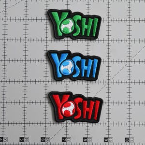 May include: Three embroidered patches featuring the word "YOSHI" in green, blue, and red, each with a matching egg design. The patches have a black border and are displayed on a gray grid background.