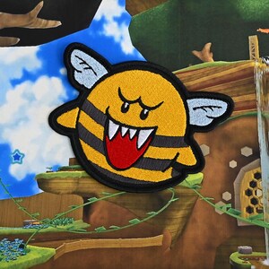Puede incluir: Un personaje amarillo y negro parecido a una abeja con una gran sonrisa de dientes y una lengua roja. El personaje tiene grandes alas blancas y est&aacute; bordado sobre un fondo de tela.
