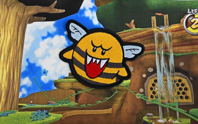 Puede incluir: Un personaje amarillo y negro parecido a una abeja con una gran sonrisa de dientes y una lengua roja. El personaje tiene grandes alas blancas y est&aacute; bordado sobre un fondo de tela.