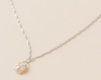 Pode incluir: Um delicado colar de prata com um único pingente de pérola de cor pêssego claro, de formato irregular. A corrente tem um design torcido e é exibida em um fundo neutro, destacando a simplicidade e elegância da joia.