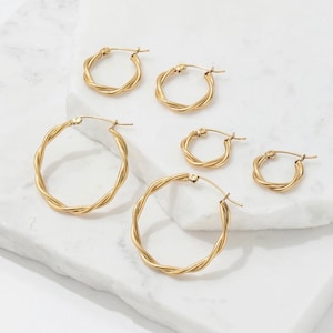 May include: Six gold-coloured twisted hoop earrings of different sizes are displayed. The earrings are arranged on a white marble surface, highlighting their design. The hoops have a classic, elegant style. The earrings are made of a gold-coloured metal.