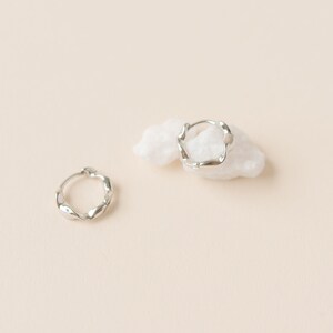 Pode incluir: Dois brincos de argola prateados com um design ondulado. Um brinco está plano, o outro repousa sobre um pequeno objeto branco em forma de nuvem. O fundo é de cor bege claro liso.