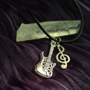 Puede incluir: Un collar con un dije de guitarra y clave de sol plateados en un cordón negro. El collar está colgado de un cristal transparente.