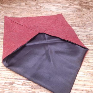 May include: A folded, square fabric item. The top layer is a deep red with a subtle, swirling pattern. The interior is a smooth, dark grey fabric. The item is displayed on a light brown, textured surface.