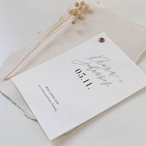 May include: A white card with black text that reads "Wir laden ein zur standesamtlichen Trauung 05.11."  The card is on a beige textured surface with a dried flower sprig.