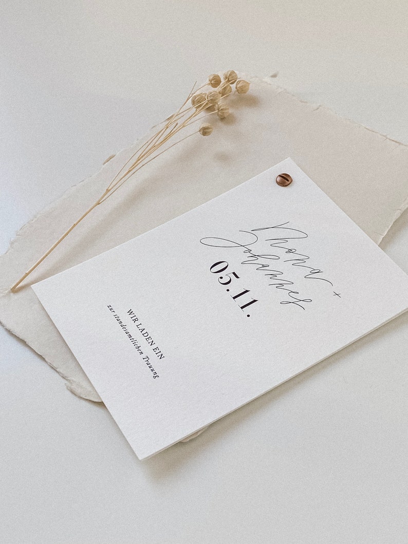 May include: A white card with black text that reads "Wir laden ein zur standesamtlichen Trauung 05.11."  The card is on a beige textured surface with a dried flower sprig.