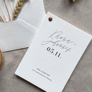 May include: A white wedding invitation with the text "Rona + John" and "05.11." in black calligraphy. The invitation also includes the text "Wir laden ein zur standesamtlichen Trauung" in black.