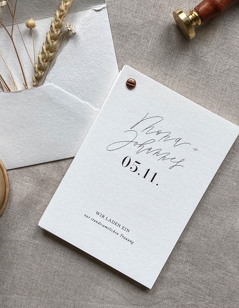 May include: A white wedding invitation with the text "Rona + John" and "05.11." in black calligraphy. The invitation also includes the text "Wir laden ein zur standesamtlichen Trauung" in black.