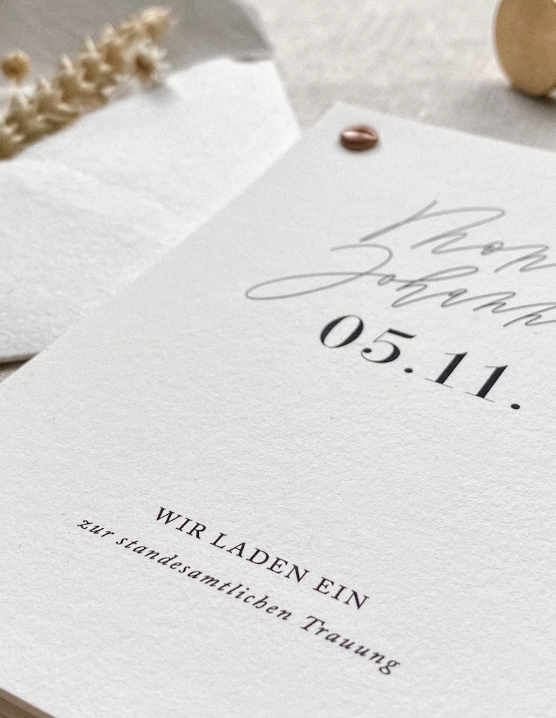 May include: A white card with black text that reads "Wir laden ein zur standesamtlichen Trauung" and "05.11."