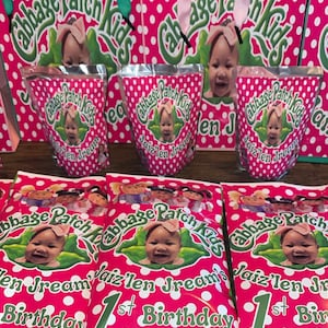 Może przedstawiać: Zestaw różowo-białych torebek i saszetek w kropki z logo Cabbage Patch Kids, zdjęciem dziecka oraz napisem Jaiz'len Jream's 1st Birthday. Produkty posiadają uchwyty wykonane z zielonej lub różowej wstążki.