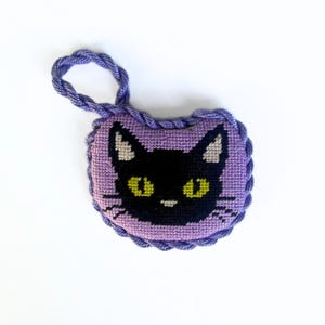 May include: A small, heart-shaped lavender cross-stitch ornament featuring a black cat face with bright yellow eyes and white inner ears. The piece is edged with a twisted purple cord that forms a loop at the top for hanging purposes.