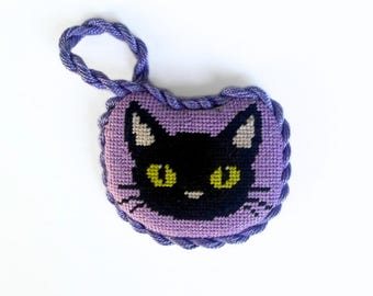 May include: A small, heart-shaped lavender cross-stitch ornament featuring a black cat face with bright yellow eyes and white inner ears. The piece is edged with a twisted purple cord that forms a loop at the top for hanging purposes.