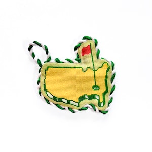 May include: A handmade golf-themed ornament in the shape of a course map. The ornament features a yellow center with a red flag, green and white rope trim, and a green border. The ornament is approximately 3 inches wide.