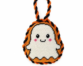 May include: A cross-stitched ornament featuring a white ghost with black eyes and pink cheeks, centered on an orange background. The ornament is framed by a twisted orange and black rope border and includes a matching twisted rope loop for hanging.
