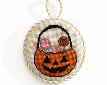 May include: A circular cross-stitch ornament featuring an orange jack-o'-lantern bucket filled with colorful round candies and lollipops. The design is stitched on cream-colored fabric and finished with a twisted beige rope border and a hanging loop.