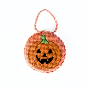 May include: A round, stuffed cross-stitch ornament featuring a jack-o'-lantern design with an orange pumpkin, a green stem, and a black face. The circular piece is edged with a twisted peach-colored cord that also forms a loop at the top.