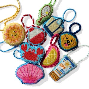 May include: A collection of colorful, hand-stitched needlepoint charms in various shapes, including a sun, crab, lemon slice, and sunscreen tube. Each charm features a loop for hanging and showcases bright colors and detailed designs, perfect for summer-themed accessories.
