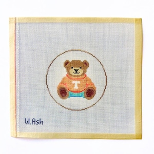 May include: Needlepoint canvas featuring a brown teddy bear wearing an orange sweater with a white "T" and blue shorts, inside a brown circle. The background is light blue, with the text "W.Ash" in the lower left corner.