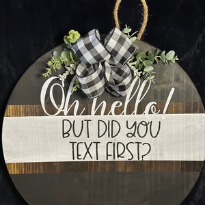 May include: A round wooden sign with a black and white checkered bow and greenery. The sign says "Oh hello! But did you text first?"