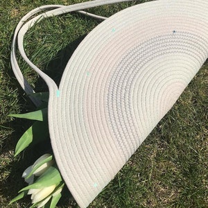 May include: A white rope tote bag with a half-circle shape and a light blue and pink ombre design. The bag has two rope handles and is sitting on a bed of green grass.