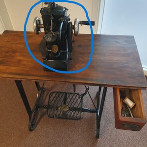 Pode incluir: Uma cabeça de máquina de costura industrial preta com uma manivela proeminente com cabo preto, uma roda de metal circular no lado esquerdo e um emblema circular dourado na carcaça frontal, projetada para tarefas de costura pesada.