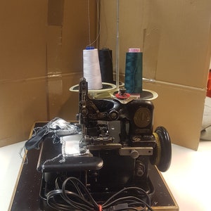 Può includere: Una macchina da cucire overlock nera con pedale e un portafilo con tre bobine di filo, una bianca, una nera e una verde.