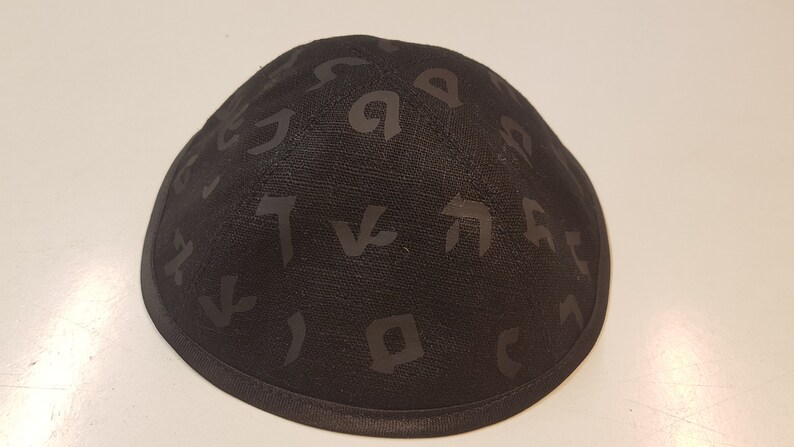 Pu&ograve; includere: Una kippah nera con un motivo di alfabeto ebraico.