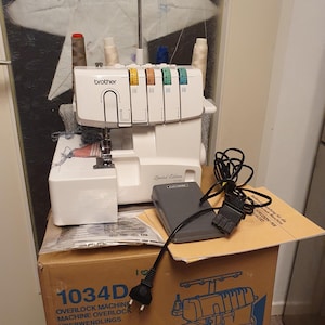 Può includere: Macchina da cucire overlock Brother 1034D bianca con più rocchetti di filo. La macchina include un pedale e si trova sopra una scatola di cartone. La scatola ha testo in più lingue.