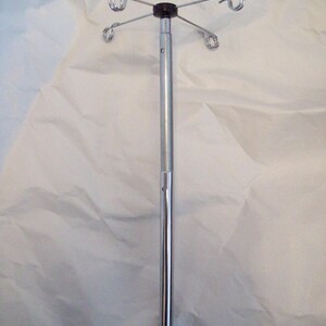 Puede incluir: Un soporte de goteo intravenoso cromado con una parte superior de plástico negro y cuatro ganchos curvos. El soporte es ajustable y tiene un poste largo y delgado. El fondo es una sábana blanca arrugada.