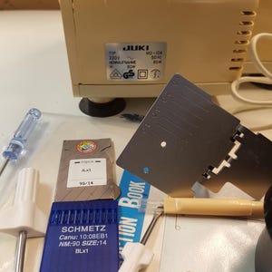 May include: A close-up of a sewing machine with a variety of sewing accessories, including needles, a screwdriver, a seam ripper, and a ruler. The sewing machine is beige and has a JUKI label with the model number MO-104. The accessories are arranged on a white surface.